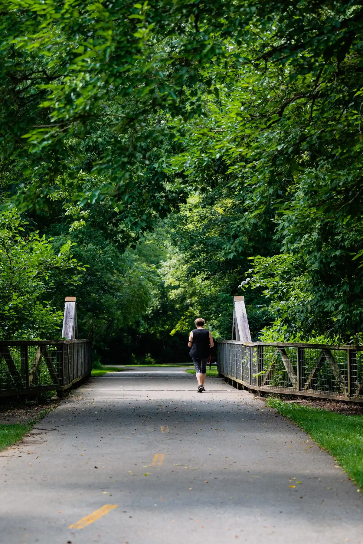 A person walks away from the camera along a paved path through a green, tree-covered tunnel, crossing a small wooden footbridge.