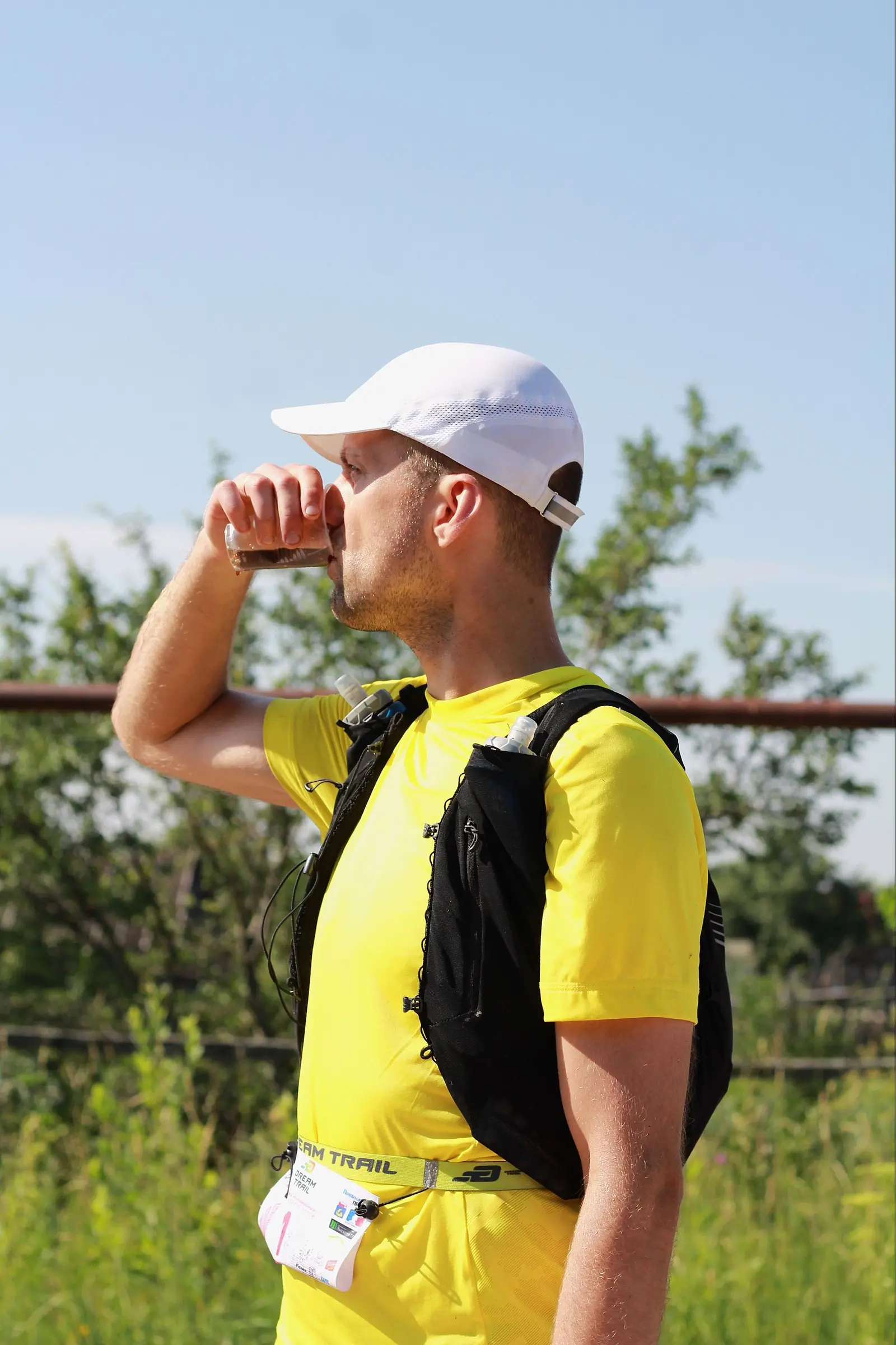 A trail runner in a yellow shirt and cap drinks from a soft flask under bright sun, sweat visible on his face, green trees in the background.